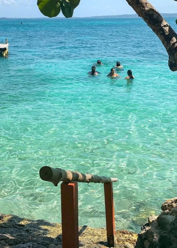 People swimming in clear turquoise water with a dock and boats in the background.