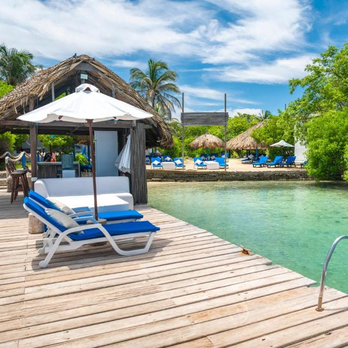 Wooden dock with lounge chairs and an umbrella overlooking a clear blue lagoon with palm trees and a thatched roof structure in the background.