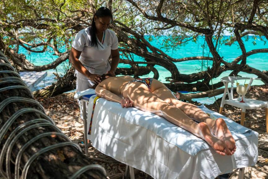 Person receiving a massage on a beach with turquoise water and trees in the background