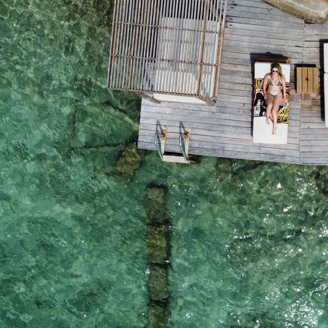Person lounging on a sunbed by a wooden deck over clear green water