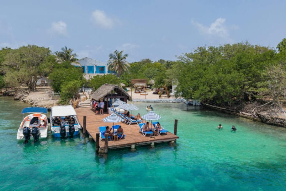 Dock with people and boats near a tropical island with clear blue water and greenery.
