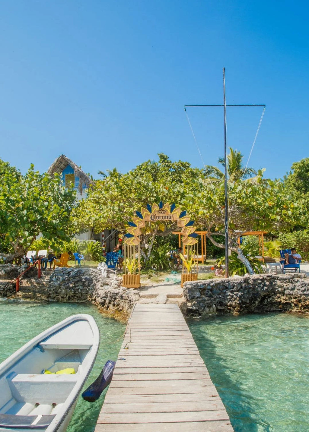 Wooden dock extending into clear blue water with a scenic background of trees and a small arch.