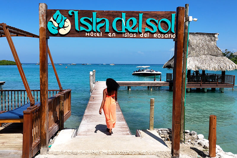 Woman walking on a dock under an 'Isla del Sol' sign with a tropical resort in the background.