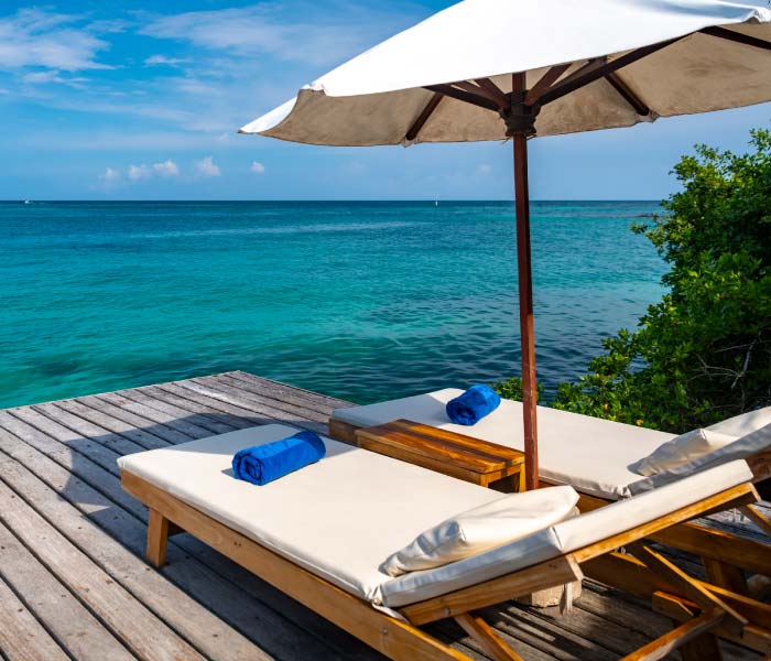 Two lounge chairs with white cushions and blue towels on a wooden deck overlooking the ocean.