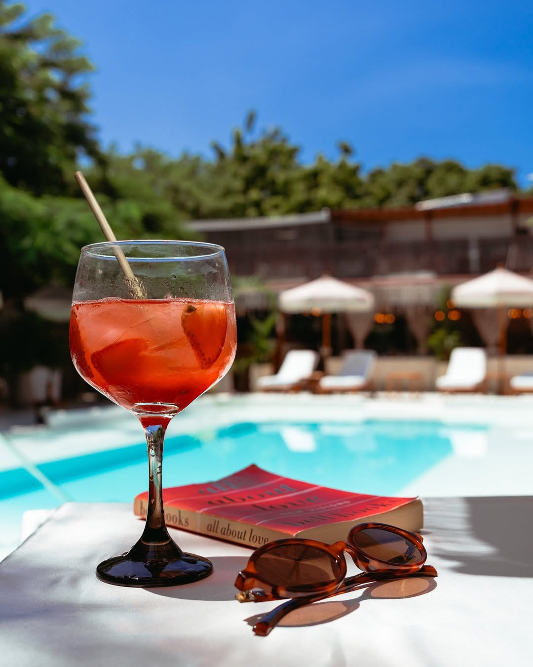 Glass of red cocktail with a straw, sunglasses, and book by a poolside with greenery and buildings in the background.