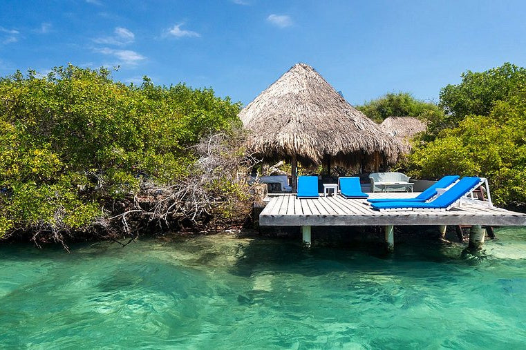Tropical setting with a thatched-roof hut and blue lounge chairs on a wooden platform over clear turquoise water.
