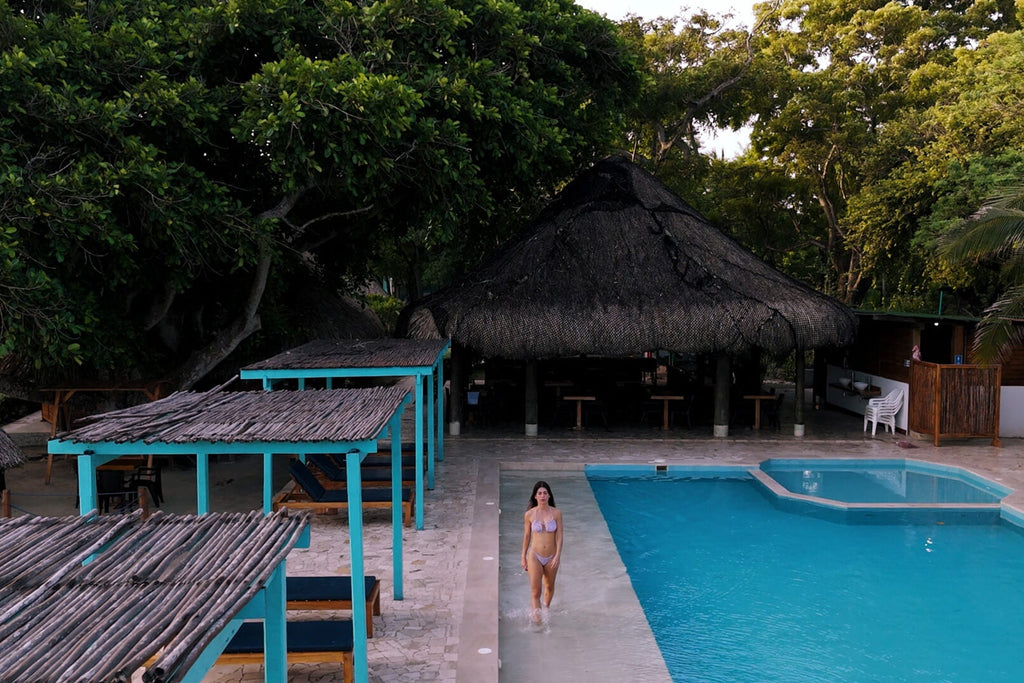 Person standing by a pool with thatched umbrellas and tables in the background