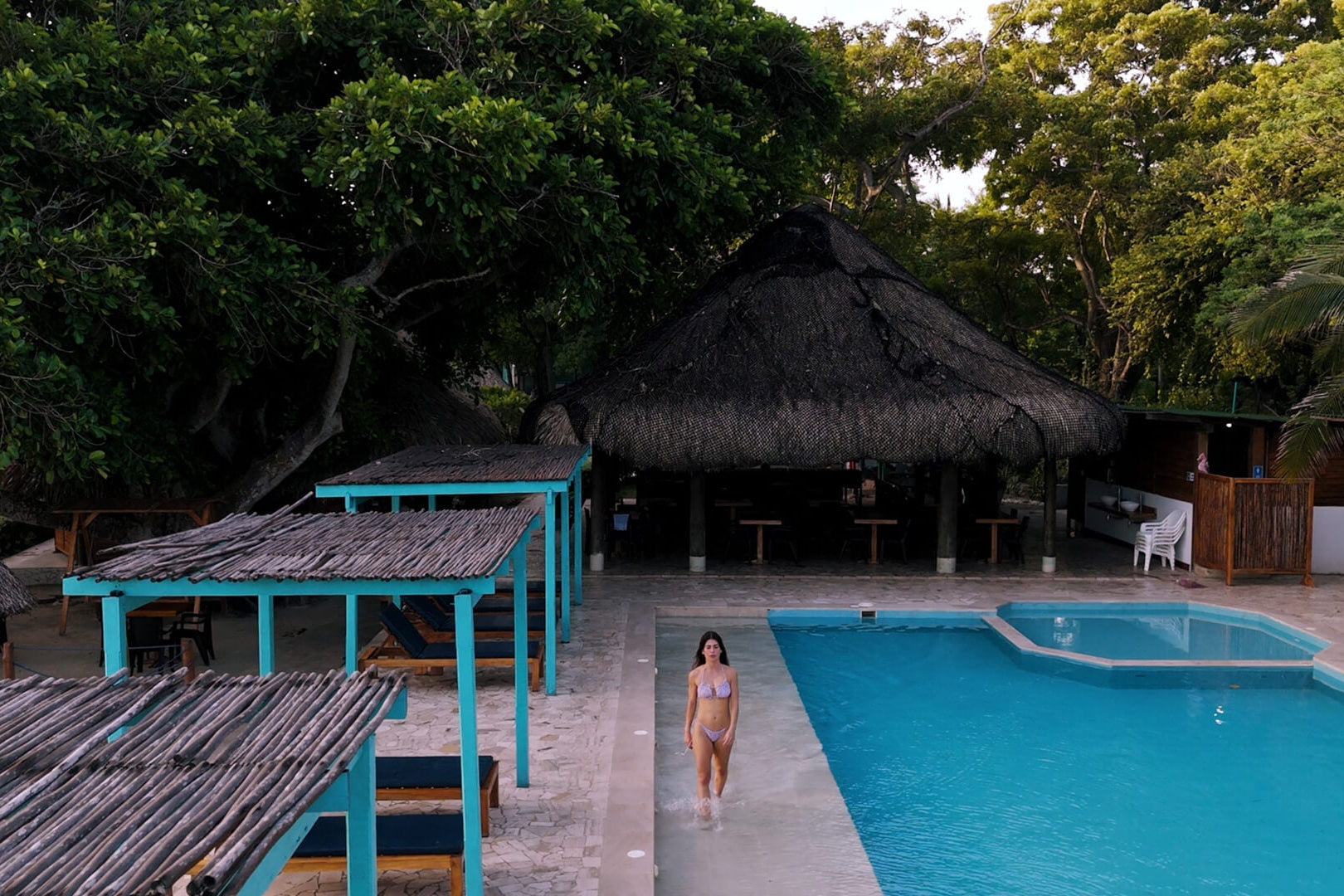 Person standing by a pool with thatched umbrellas and tables in the background