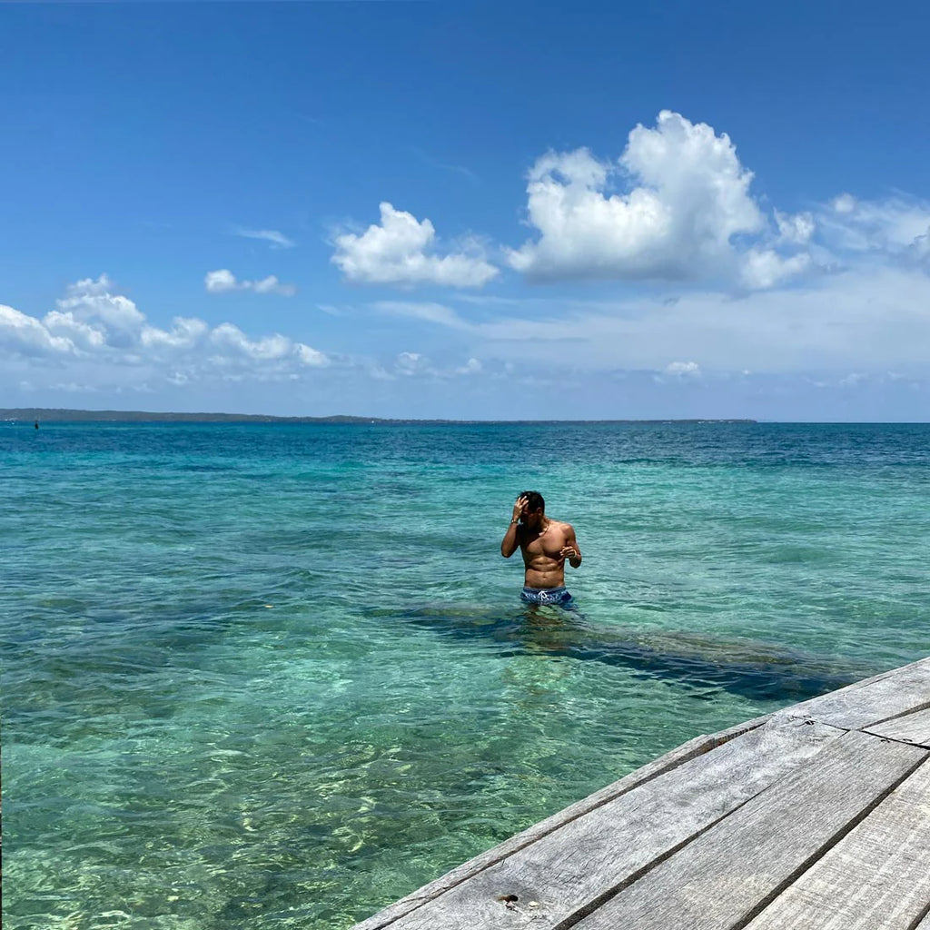 Person standing in clear blue water near a wooden dock with a blue sky and clouds in the background.