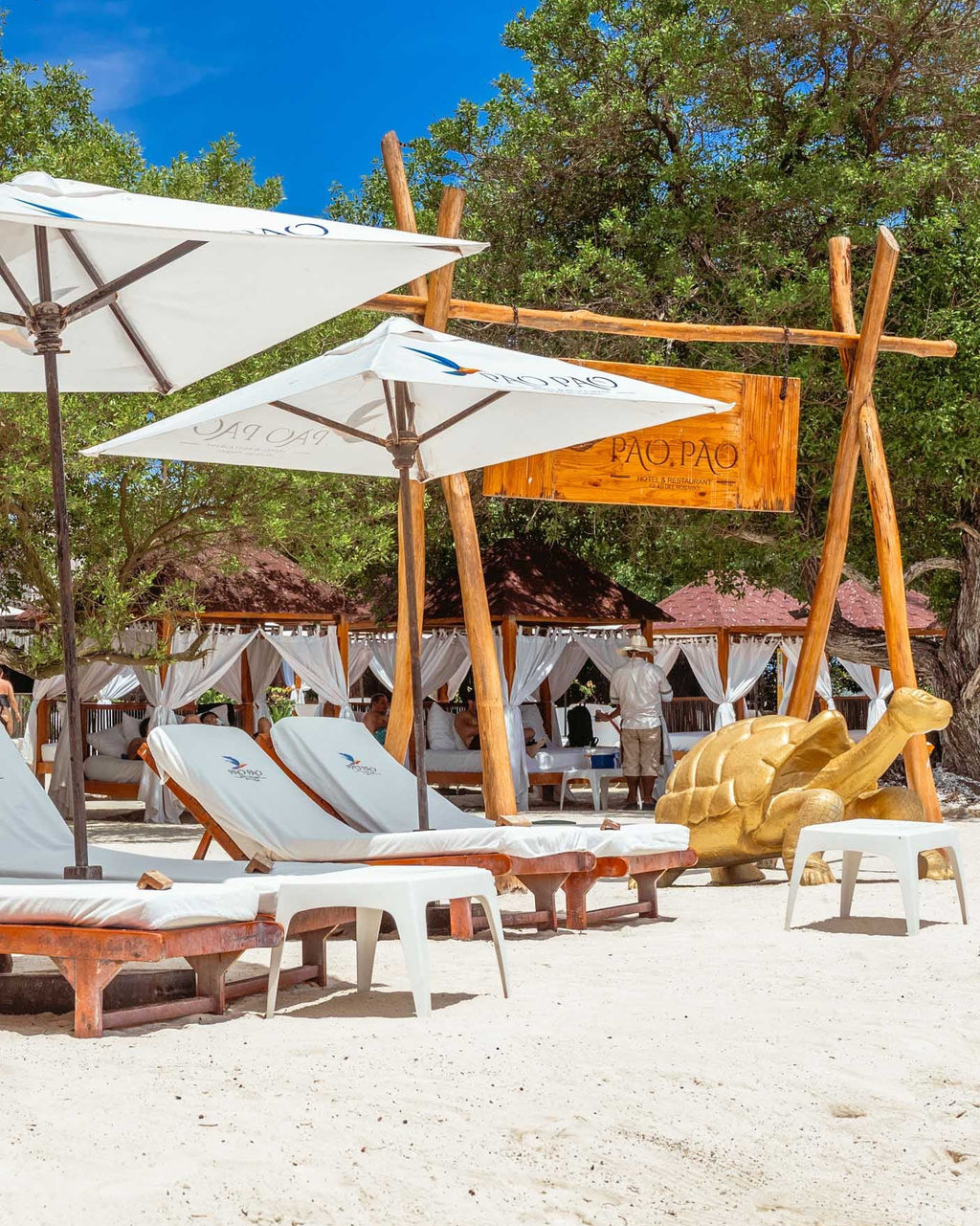 Beachside relaxation area with lounge chairs, umbrellas, and a wooden sculpture on a sandy beach.